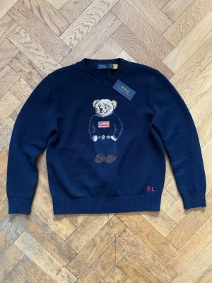 Mörkblå stickad tröja Polo Bear - Mörkblå stickad tröja från Polo Ralph Lauren med ikoniska Polo Bear-motivet på bröstet. Tröjan har rund hals, ribbstickade muddar och nederkant samt broderade initialer 'RL' i rött vid nederkanten. Perfekt för dig som gillar klassisk preppy stil. Säljer eftersom att den inte passar mig.