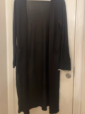 Svart lång kappa från H&M - Stilren svart lång kappa från H&M med öppen framsida och raka ärmar. Enkel design utan synliga knappar eller dragkedja, perfekt för lager på lager. Tillverkad i ett mjukt och lätt material som ger ett snyggt fall.