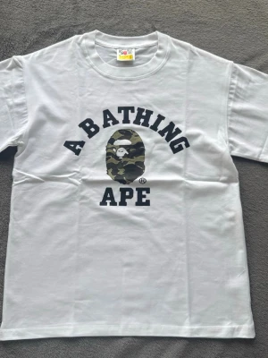  BAPE t-shirt  - Vit t-shirt från A Bathing Ape med klassisk svart text och den ikoniska ap-loggan i camouflage på bröstet. T-shirten har rund hals och korta ärmar, perfekt för en streetwear-look. Materialet är mjuk bomull. Oanvänd. Skriv vid frågor!!