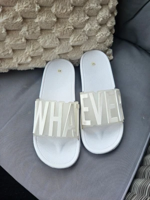 Vita sandaler med texten WHATEVER - Coola vita sandaler med bred rem där det står 'WHATEVER' i stora 3D-bokstäver. Sandalerna har en klassisk slip-in design och är perfekta på väg till poolen eller stranden. Perfekta för en avslappnad och trendig stil.