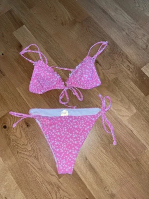 Rosa blommig bikini från 157 - Supersöt rosa bikini från 157 med vita blommor. Både överdel och underdel har knytband i sidorna och toppen har trekantsform. Perfekt för strandhäng eller poolparty i sommar!