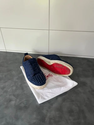 Blå sneakers med nitar från Louboutin - Snygga blå sneakers från Christian Louboutin i mocka med ikoniska röda sulor och coola nitar på tån. Skorna har svarta skosnören och en vit sula som ger en schysst kontrast. Perfekta för dig som vill sticka ut med din stil. Tyvärr finns det inte alla nitar på men det är inget som man tänker på när dem sitter på, man kan även se de på bilderna. 