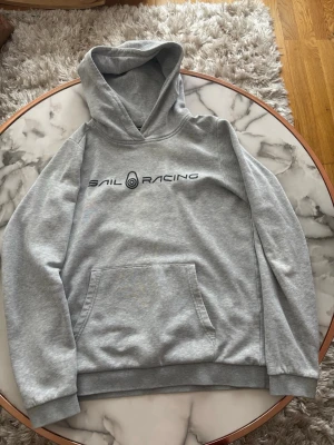 Grå hoodie från Sail Racing - Snygg grå hoodie från Sail Racing med svart logga tryckt på bröstet. Klassisk känguruficka framtill och mjuk insida. Perfekt för chill dagar eller när du vill ha en enkel och stilren look. Tillverkad i mjukt bomullsmaterial.