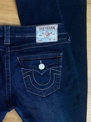 Mörkblå bootcut jeans True Religion - Säljer pågrund av att jag upplever de som stora i storleken.  På lappen står det W26 men jag tycker att de känns mer som ungefär W28, vilket motsvarar typ S-M.  Innerbens längden= ca 81 cm, ytterbens längden= ca 103 cm,  midjan (tvärs över) = ca 39/40 cm  Inga synliga defekter förutom något slitna längst ned på benet och lite urtvättade i färgen.  Modellen= Joey low rise flare. 