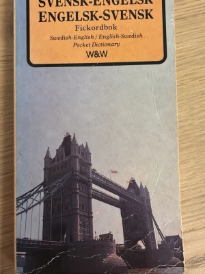 Svensk-Engelsk Fickordbok W&W - Praktisk fickordbok för svensk-engelsk och engelsk-svensk översättning. Omslaget är ljusgrått med en bild på Tower Bridge och har en orange etikett med titel och förlag. Perfekt att ha med sig i väskan när du pluggar språk eller reser.