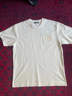 Cream DG t-shirt från Dolce & Gabbana - Säljer en stilren vit t-shirt från Dolce & Gabbana med upphöjt DG-tryck på bröstet. Klassisk rund halsringning och korta ärmar. Tillverkad i mjuk bomull för en bekväm känsla och clean look. Heavy t shirt.