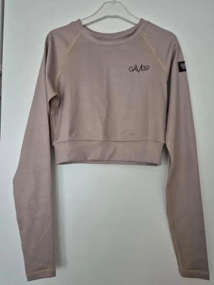 Croppad tröja - Snygg beige cropped tröja från Gavelo med lång ärm och rund halsringning. Tröjan har en stilren design med logga på bröstet och patch på ärmen. Perfekt till jeans eller träningstights. Trendig, kortärmad kort tröja från Gavelo. Perfekt för både träning och vardag. Den beige färgen gör den lätt att matcha med andra plagg.