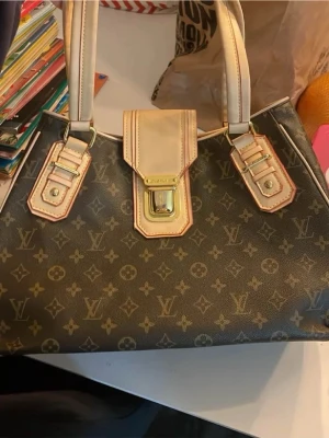 Monogram handväska - handväska i brunt monogramcanvas med beige läderdetaljer och guldiga metalldelar. Väskan har två rejäla handtag och ett lock med spänne framtill. Ikoniskt mönster över hela väskan och snygga kontrastsömmar. Lite ”sprucken” i materialet på beiga knapptygey se bild nummer 2 