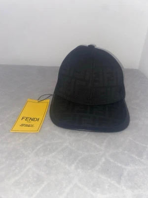 Svart  Fendi  keps med logomönster - Snygg svart Fendi keps  med diskret FF-logomönster över hela Fendi  kepsen. Klassisk böjd skärm och justerbar rem baktill med gulddetalj. Tillverkad i textilmaterial som ger en lyxig känsla och stilren look.