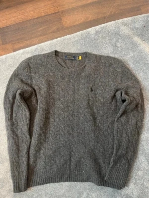 Grå kabelstickad tröja Polo Ralph Lauren - Mysig grå kabelstickad tröja från Polo Ralph Lauren med rund halsringning och diskret broderad logga på bröstet. Tröjan har långa ärmar och ribbade muddar vid ärmslut och nederkant. Perfekt för kyliga dagar och enkel att matcha.