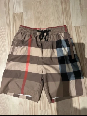 Beige rutiga shorts med snörning - Snygga beige shorts från Burberry med klassiskt rutigt mönster i brunt, svart, rött och vitt. Shortsen har elastisk midja med svart snörning och sidofickor. Perfekta för sommaren och ger en trendig look.
