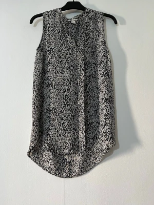 Svartvit blus med leopardmönster - Snygg ärmlös blus i svartvitt leopardmönster. Modellen är rak med rund halsringning och lätt rundad nederkant. Perfekt för dig som gillar trendiga och bekväma toppar med lite extra edge.