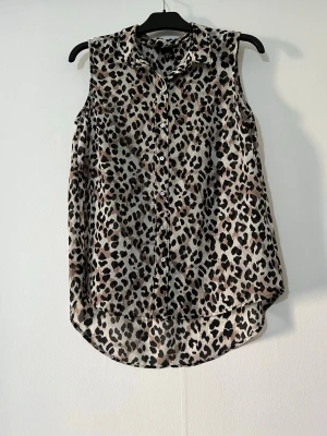 Ärmlös leopardmönstrad blus - Snygg ärmlös blus med leopardmönster i beige, brunt och svart. Blusen har krage, knäppning framtill och rundad nederkant som är längre bak. Tillverkad i ett lätt och luftigt material som passar perfekt till sommaren.