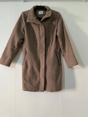 Beige kappa med dragkedjor  - Stilren beige kappa med hög krage och två sneda fickor med dragkedjor. Kappan har lång ärm och stängs med dold knäppning framtill. Perfekt för kyliga dagar och ger en clean look till din outfit. Köpt från Cassels