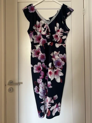 Blommig klänning med volang - Snygg marinblå klänning med stora blommor i rosa och vitt. Klänningen har v-ringning, volangdetalj vid axlarna och är knälång. Perfekt för dig som vill sticka ut med ett färgstarkt mönster. Materialet känns mjukt och följsamt.