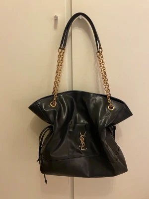 Svart handväska från YSL med guldkedja - Svart YSL handväska i mjukt läder med guldfärgade kedjedetaljer och dragsko på sidorna. Rymlig modell i nyskick.