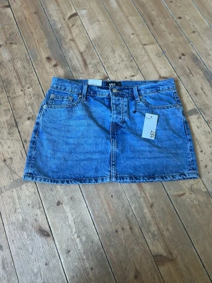 Blå jeanskjol från 157 - Snygg blå jeanskjol från 157 med knappar framtill. Kjolen har en rak passform. Passar till många olika outfits och är ett måste i garderoben.