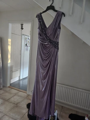 Lila långklänning med paljettdetaljer - Elegant lila långklänning med draperad design och v-ringning. Klänningen har en axelbandslös look på ena sidan och är dekorerad med glittrande paljetter längs midjan. Tillverkad i ett glansigt material som ger en lyxig känsla. Storlek 42☺️