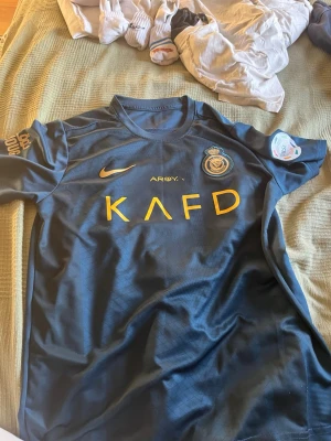 Al Nassr Ronaldo matchtröja blå/guld - Snygg Al Nassr matchtröja i mörkblått med guldiga detaljer och tryck. Framsidan har klubbmärke, Nike-logga och sponsortryck, medan baksidan har 'Ronaldo' och nummer 7 i guld. Perfekt för fotbollsfans som vill sticka ut på planen eller läktaren.