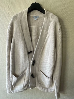Beige cardigan från DKNY med knappar - Snygg beige stickad cardigan från DKNY. Koftan har bred ribbad krage och muddar vid ärmslut och nederkant. Perfekt för lager-på-lager och enkel att matcha med olika outfits.