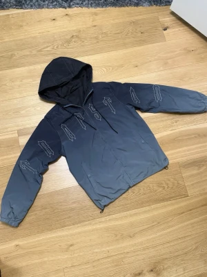 Trapstar jacka -  Grå vindjacka med huva och dragkedja framtill. Jackan har vita grafiska tryck på bröstet och ärmarna, samt justerbar dragsko i nederkant. Tillverkad i lätt och smidigt material, perfekt för blåsiga dagar.