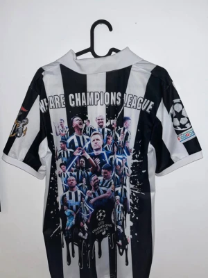 Newcastle United Champions League tröja - Snygg Newcastle United matchtröja i svart och vitt med Champions League-tema. Framsidan har klubbmärke, Castore-logga och sponsortryck. Baksidan har stort tryck med spelare och texten 'We Are Champions League'. Perfekt för fotbollsfans!