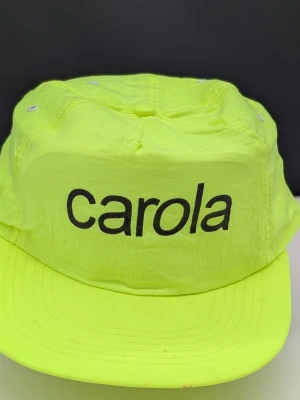 Neongul keps med svart Carola Häggvist tryck - Cool neongul keps med svart tryckt Carola' framtill. Kepsen har klassisk form med böjd skärm och ventilationshål på ovansidan. Tillverkad i lätt och tunt material som ger en sportig vibe. Officiell merchandise från tiden det begav sig 
