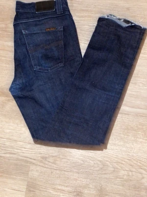 Mörkblå jeans från Nudie Jeans - Säljer ett par mörkblå jeans från Nudie Jeans i klassisk rak modell. Jeansen har fem fickor, tydliga sömmar och rå kant vid benslutet för en trendig look. Tillverkade i denim av bomull. Perfekta för dig som gillar stilrena och tidlösa jeans. Kom med frågor eller prisförslag🤝😇