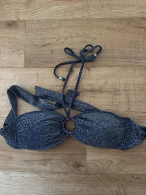 Glittrig mörkblå bikiniöverdel - Snygg glittrig mörkblå bikiniöverdel med vadderade kupor och knyt i nacken. Bikinin har en liten ringdetalj framtill och smala axelband med silverfärgade detaljer. Perfekt för strandhäng eller poolparty.