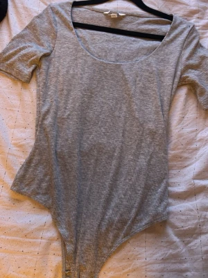 Grå ribbad body med kort ärm - En enkel och stilren grå body med ribbad struktur och korta ärmar. Bodyn har en djup rundad urringning och knäppning i grenen. Perfekt att matcha med jeans eller kjol för en clean look.