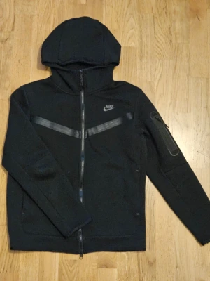 Svart Nike hoodie med dragkedja - Snygg svart hoodie från Nike med hel dragkedja framtill och huva. Har två svarta reflexdetaljer över bröstet och en ficka med dragkedja på ena ärmen. Perfekt för dig som gillar sportig och clean stil. Nike-logga på bröstet och mjukt material.