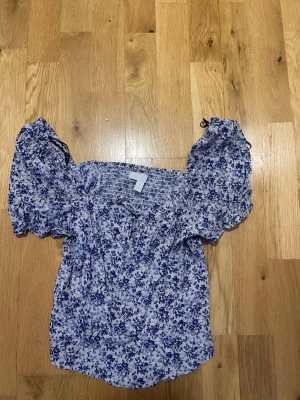 HM Blommig offshoulder blus med puffärm stl S - Supersöt blommig blus i vitt och blått med offshoulder-modell och puffiga korta ärmar. Smockad upptill för en bekväm passform och voluminösa ärmar som ger en romantisk vibe. Perfekt till jeans eller kjol.