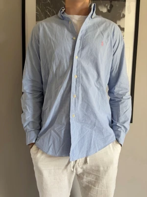 Blå skjorta från Ralph Lauren - Klassisk blå skjorta från Ralph Lauren med diskret rutigt mönster och broderad logga i rosa på bröstet. Skjortan har lång ärm, knappar framtill och en normal passform. Perfekt för en stilren look.