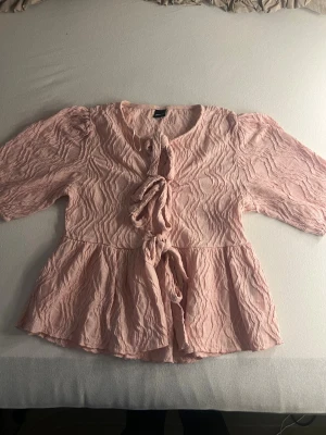 Rosa blus med knyt och struktur - Söt ljusrosa blus från Gina med knyt framtill och volang nertill. Blusen har en crinklad struktur i tyget och trekvartsärmar med lätt puff. Perfekt för en trendig och feminin look.❤️