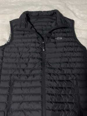 Svart dunväst från The North Face - Snygg svart dunväst från The North Face med quiltad design och hög krage. Västen har dragkedja framtill och två sidofickor med dragkedja. Perfekt för lager-på-lager och håller dig varm under kyliga dagar.