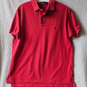 Röd pikétröja från Polo Ralph Lauren - Klassisk röd pikétröja från Polo Ralph Lauren med korta ärmar och krage. Tröjan har knappar vid halsen och den ikoniska broderade loggan på bröstet. Perfekt för en stilren och avslappnad look.