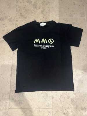 Svart Maison Margiela t-shirt - Cool svart t-shirt från Maison Margiela med vit och grön logga framtill. Klassisk passform och rund halsringning. Tillverkad i mjuk bomull, perfekt för en trendig och avslappnad stil.
