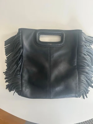 Maje väska - Cool svart handväska i läder från Maje med fransar på sidorna. Väskan har en avtagbar axelrem och kommer med en dustbag (se bild 2). Så gott som nytt skick förutom att märket på väskan skrapats bort jättelite (bild 3.) Kan tänka mig att diskutera pris något vid snabb affär. Skriv vid fler  frågor och funderingar.💞
