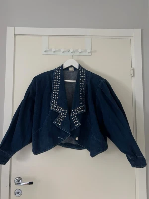 Oversized jeansjacka med nitar - Mörkblå oversized jeansjacka med bred krage dekorerad med silverfärgade nitar. Jackan har en kortare, bolero liknande passform samt axelvaddar, men som går att klippa bort. Står ej storlek men sitter lite oversized på S