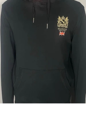Svart hoodie från Morris - Svart hoodie från Morris med guldfärgat tryck och brittisk flagga på bröstet. Klassisk känguruficka framtill och justerbar huva med snören. Tillverkad i mjukt material som passar perfekt för en avslappnad stil. Skriv för ett prisförslag🙌