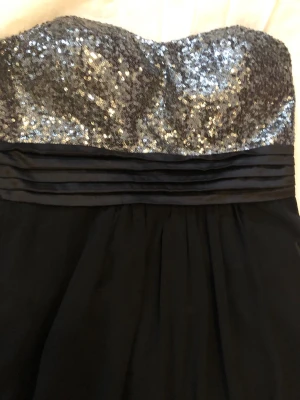 Svart långklänning med paljetter - Elegant svart långklänning med axelbandslös design och glittrig överdel täckt av silverfärgade paljetter. Kjolen är lång och faller mjukt, medan midjan har veckade detaljer. Perfekt för bal eller festliga tillfällen.