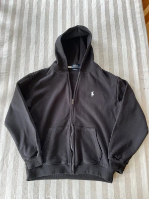 Svart hoodie från Polo Ralph Lauren - Svart hoodie med dragkedja från Polo Ralph Lauren. Klassisk modell med huva, två fickor framtill och broderad vit logga på bröstet. Mjuk bomullsblandning och ribbade muddar vid ärmslut och nederkant. Perfekt för en avslappnad och stilren look.