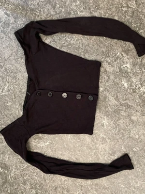 Svart ribbad kofta med knappar - Snygg svart ribbad kofta med långa ärmar och fem stora knappar framtill. Kortare modell som sitter tajt och har en modern, croppad passform. Perfekt att styla över ett linne eller t-shirt för en trendig look.