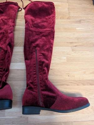 Vinröda overknee-stövlar i sammet - Snygga vinröda overknee-stövlar i sammetsmaterial med låg klack och dragkedja på sidan. Stövlarna har rund tå och snörning upptill för bättre passform. Perfekta för att lyfta din outfit med en trendig touch.