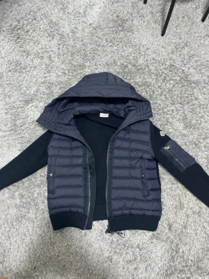 Mörkblå Moncler cardigan  - Säljer nästan helt ny inga defekter jätte Snygg mörkblå pufferjacka från Moncler med stickade ärmar och huva. Jackan har dragkedja framtill, två fickor med knappar och en extra ficka med dragkedja på ärmen. Klassisk Moncler-logga på armen och quiltad design framtill.