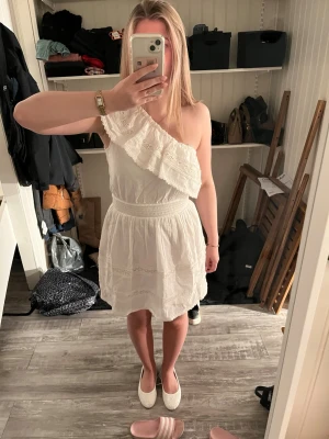 Vit oneshoulder klänning med volang - Söt vit klänning i oneshoulder-modell med bred volang och spetsdetaljer. Klänningen har resår i midjan och är tillverkad i ett lätt bomullstyg med diskret struktur. Perfekt för sommarens alla tillfällen och studenten! Helt ny, nypris 629.