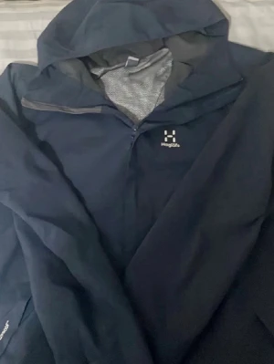 Marinblå vindjacka från Haglöfs - Säljer en svart vindjacka från Haglöfs med dragkedja och hög krage. Jackan har en minimalistisk design med diskret logga på bröstet och är tillverkad i ett lätt och slitstarkt material. Perfekt för blåsiga dagar och enkel att bära med sig.