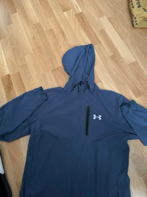 Blå vindjacka från Under Armour - Snygg blå vindjacka från Under Armour med huva och dragkedja framtill. Jackan har en bröstficka med dragkedja och logga på vänster sida. Tillverkad i lätt och tåligt material, perfekt för blåsiga dagar.