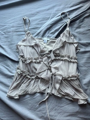 Beige volangtop från Misu - Supersöt beige linnetopp från Misu med volanger och knyt framtill. Toppen har tunna axelband och är tillverkad i ett lätt, plisserat material som ger en flowy känsla. Perfekt att styla med jeans eller kjol för en trendig look.