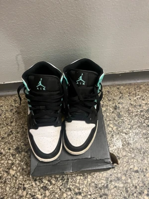 Air Jordan 1 Mid turkos och svart - Snygga Air Jordan 1 Mid sneakers i svart, vitt och turkos. Skorna har klassisk Jordan-logga på plösen, svarta skosnören och perforerad tå. Ovandelen är i läder och textil, med turkosa detaljer runt hälen och ankeln. Perfekt för dig som gillar streetwear och basketstil.